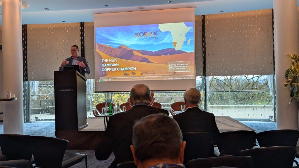 Male presenter at a podium giving a talk; slide reads 'The New Namibian Copper Champion' with desert dunes and an Africa map in the background.