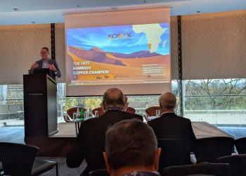Male presenter at a podium giving a talk; slide reads 'The New Namibian Copper Champion' with desert dunes and an Africa map in the background.