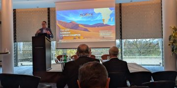 Male presenter at a podium giving a talk; slide reads 'The New Namibian Copper Champion' with desert dunes and an Africa map in the background.