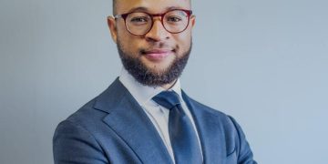 Zach Kauraisa | Mining and Energy Namibia | Namibia’s Leading Mining & Energy News Professional portrait: man in a navy suit and tie, wearing red glasses, smiling at the camera.