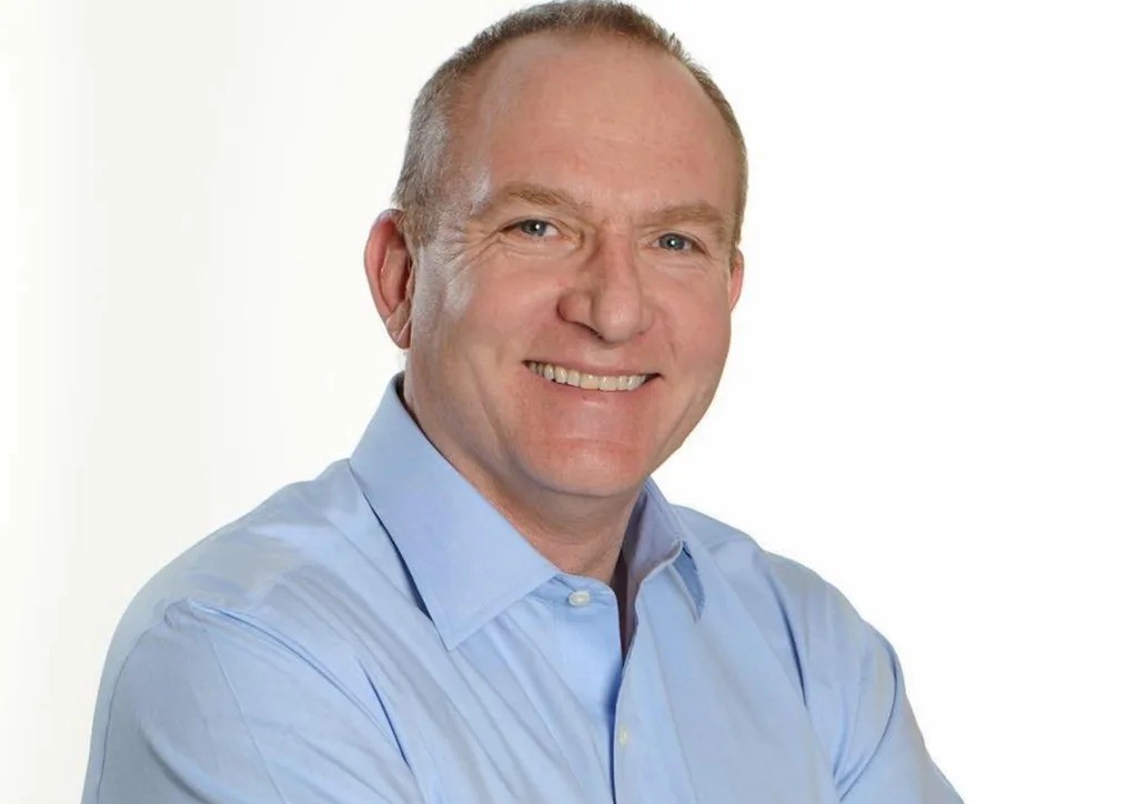 Smiling middle-aged man in a light blue dress shirt, head-and-shoulders portrait against a white background.