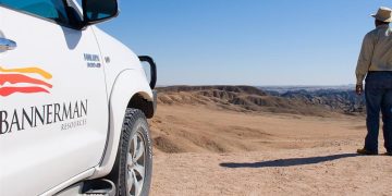 White pickup truck with Bannerman Resources branding in the desert, a man in a hat stands at the edge looking over arid hills under a blue sky.