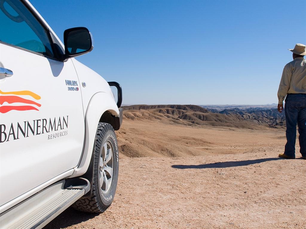 Bannerman Energy | Mining and Energy Namibia | Namibia’s Leading Mining & Energy News White pickup truck with Bannerman Resources branding in the desert, a man in a hat stands at the edge looking over arid hills under a blue sky.