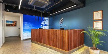 Reception desk in a modern office lobby with a blue wall and company logo, wood-panel front, and a potted plant nearby.