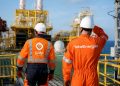 Two workers in bright orange safety suits and white hard hats stand on an offshore oil platform, surveying equipment.