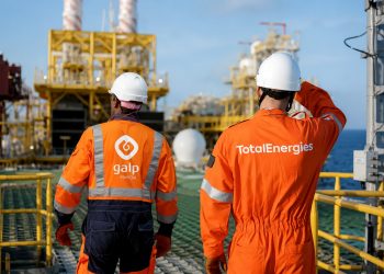 total energies | Mining and Energy Namibia | Namibia’s Leading Mining & Energy News Two workers in bright orange safety suits and white hard hats stand on an offshore oil platform, surveying equipment.
