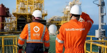 Two workers in bright orange safety suits and white hard hats stand on an offshore oil platform, surveying equipment.