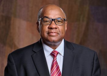 JOHANNES GAWAXAB 1 | Mining and Energy Namibia | Namibia’s Leading Mining & Energy News Professional headshot of an older man in a dark suit and red striped tie, wearing glasses against a brown backdrop.
