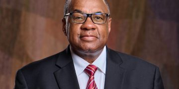 Professional headshot of an older man in a dark suit and red striped tie, wearing glasses against a brown backdrop.