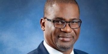 Eugene Okpere | Mining and Energy Namibia | Namibia’s Leading Mining & Energy News Professional headshot of a man wearing a navy suit and blue tie, glasses, against a blue studio backdrop.