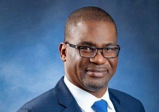 Professional headshot of a man wearing a navy suit and blue tie, glasses, against a blue studio backdrop.