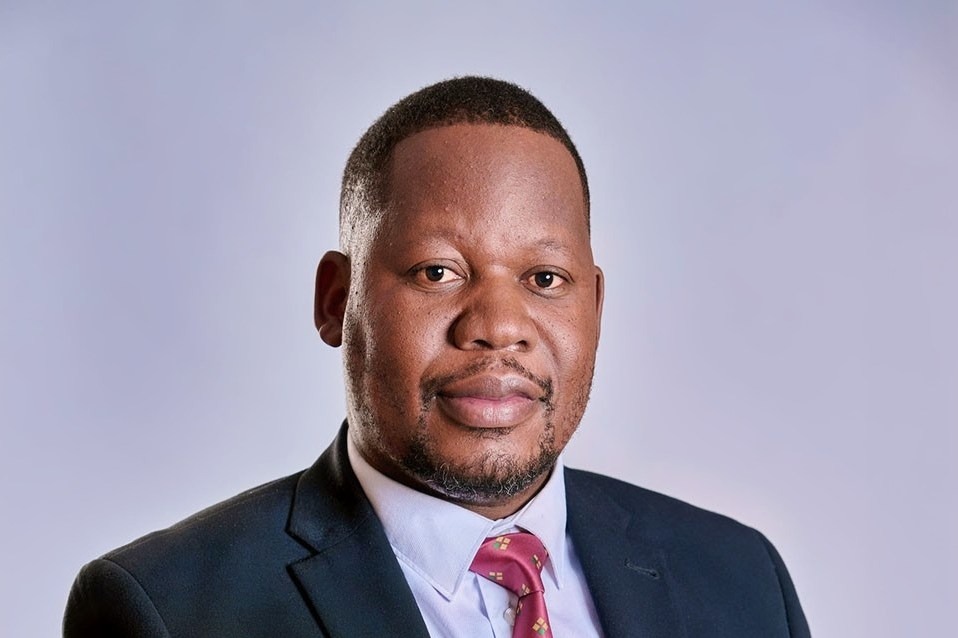 Nicholas Mukasa | Mining and Energy Namibia | Namibia’s Leading Mining & Energy News Professional headshot of a man in a dark suit, light blue shirt, and red tie against a neutral gray background.