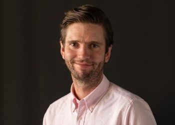 Portrait of a man with light beard wearing a pink button-down shirt, smiling against a dark background.