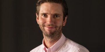 Portrait of a man with light beard wearing a pink button-down shirt, smiling against a dark background.