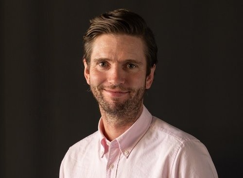 Gerard ODonovan | Mining and Energy Namibia | Namibia’s Leading Mining & Energy News Portrait of a man with light beard wearing a pink button-down shirt, smiling against a dark background.