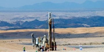 Oil drilling rig in a barren desert, workers in hi-vis near machinery with mountains in the distance.