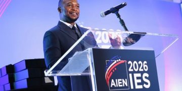 Male presenter in a suit speaks at a clear podium on a lit stage; sign reads '2026 IES' and 'New Orleans' in the foreground.