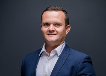 John Roos - Corporate Headshots | 2025 | Mining and Energy Namibia | Namibia’s Leading Mining & Energy News Head-and-shoulders portrait of a smiling man in a blue suit and white shirt against a dark gray backdrop.