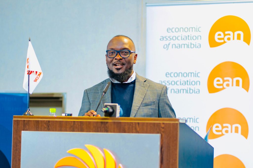 KASUTO | Mining and Energy Namibia | Namibia’s Leading Mining & Energy News Male speaker at a podium during an Economic Association of Namibia event, wearing glasses and a gray blazer with a banner in the background showing 'economic association of namibia' and 'ean' logos.