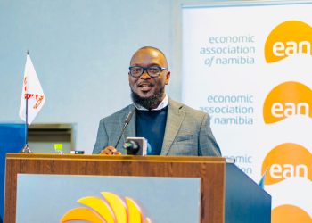 Male speaker at a podium during an Economic Association of Namibia event, wearing glasses and a gray blazer with a banner in the background showing 'economic association of namibia' and 'ean' logos.