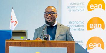 Male speaker at a podium during an Economic Association of Namibia event, wearing glasses and a gray blazer with a banner in the background showing 'economic association of namibia' and 'ean' logos.