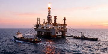 Offshore oil rig with a tall flare stack at sunset, connected to two escort ships in calm sea.