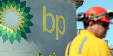 BP logo on a large cylindrical storage tank with a worker in yellow safety gear in the foreground against a blue sky.