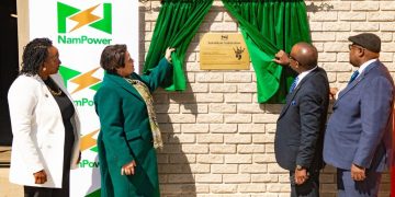 Four officials unveil a bronze plaque on a brick wall while green curtains frame it, next to a NamPower banner.