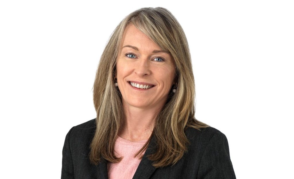 Professional headshot of a smiling woman in a dark blazer and pink top against a white background.
