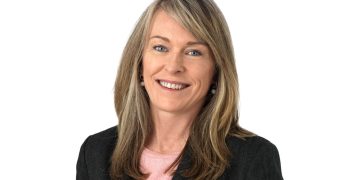 Professional headshot of a smiling woman in a dark blazer and pink top against a white background.