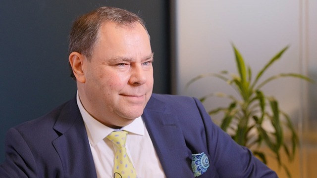 Jeremy Asher | Mining and Energy Namibia | Namibia’s Leading Mining & Energy News Man in a navy suit and yellow patterned tie sits in an office, with a plant in the background and a calm, thoughtful expression.