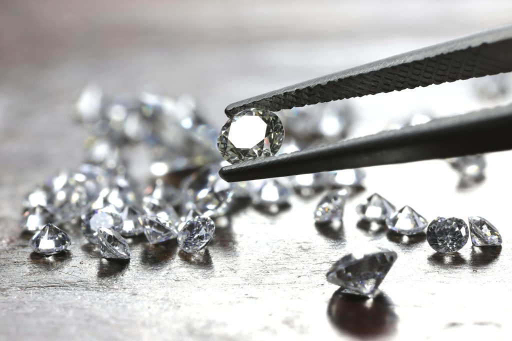 Round-cut diamond held by tweezers with loose diamonds scattered on a wooden surface in the background.