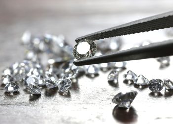 Round-cut diamond held by tweezers with loose diamonds scattered on a wooden surface in the background.