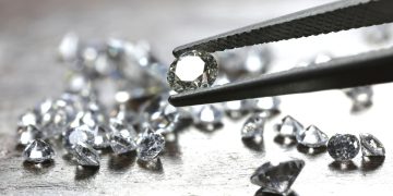 diamond 7 | Mining and Energy Namibia | Namibia’s Leading Mining & Energy News Round-cut diamond held by tweezers with loose diamonds scattered on a wooden surface in the background.