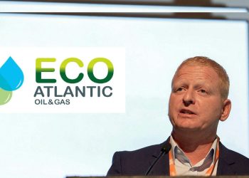 Man in a suit speaking at a podium during a conference, with the ECO Atlantic Oil & Gas logo on screen behind him, and a microphone in front.