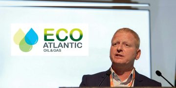 Gil Holzman | Mining and Energy Namibia | Namibia’s Leading Mining & Energy News Man in a suit speaking at a podium during a conference, with the ECO Atlantic Oil & Gas logo on screen behind him, and a microphone in front.