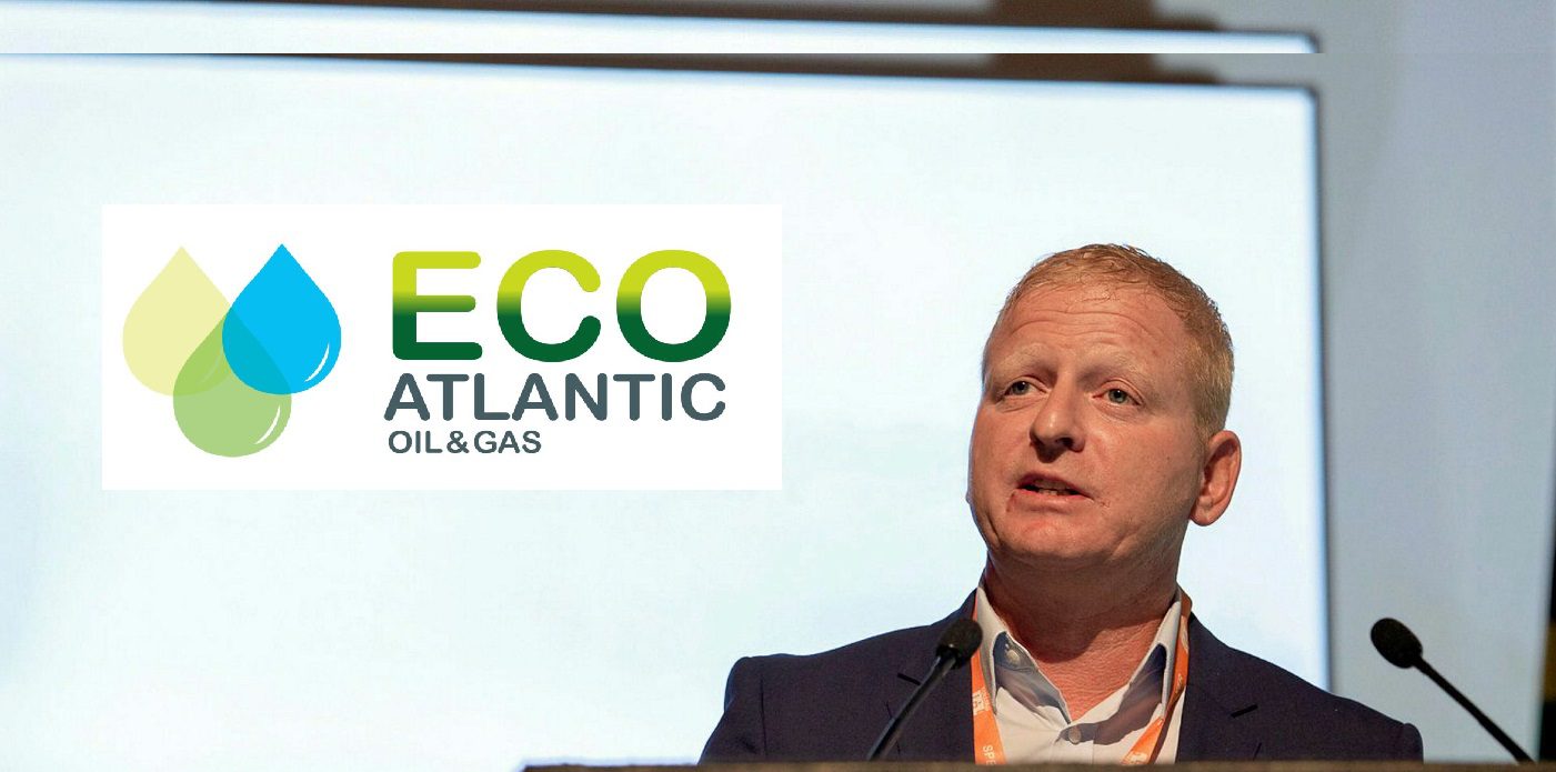 Man in a suit speaking at a podium during a conference, with the ECO Atlantic Oil & Gas logo on screen behind him, and a microphone in front.