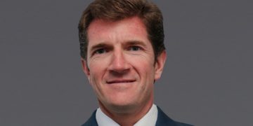 Portrait of a middle-aged man in a navy suit and light blue tie against a gray background, smiling subtly.
