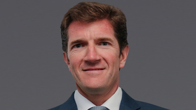 Portrait of a middle-aged man in a navy suit and light blue tie against a gray background, smiling subtly.