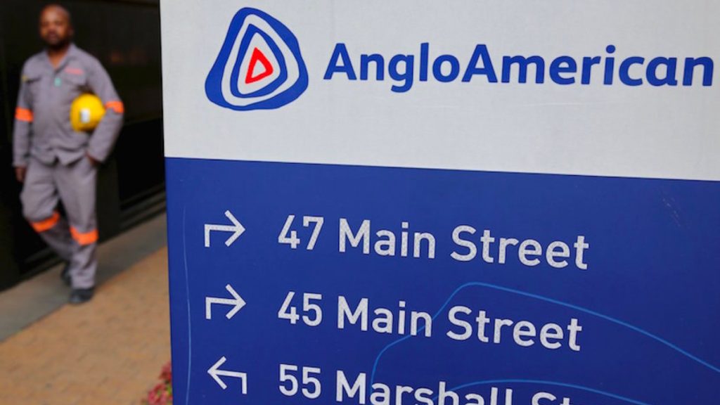 Signboard with AngloAmerican logo and arrows to 47 Main Street, 45 Main Street, and 55 Marshall Street; worker in gray uniform holding a yellow helmet in the background.