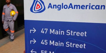 Signboard with AngloAmerican logo and arrows to 47 Main Street, 45 Main Street, and 55 Marshall Street; worker in gray uniform holding a yellow helmet in the background.