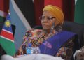 Portrait of an elderly woman wearing a yellow head wrap and a colorful patterned dress, seated at a conference with the Namibian flag in the background.