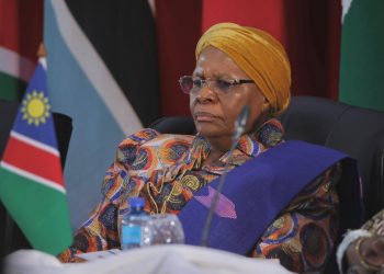 NAMIBIA PRESIDENT | Mining and Energy Namibia | Namibia’s Leading Mining & Energy News Portrait of an elderly woman wearing a yellow head wrap and a colorful patterned dress, seated at a conference with the Namibian flag in the background.