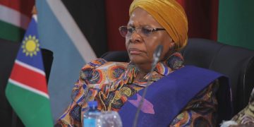 Portrait of an elderly woman wearing a yellow head wrap and a colorful patterned dress, seated at a conference with the Namibian flag in the background.