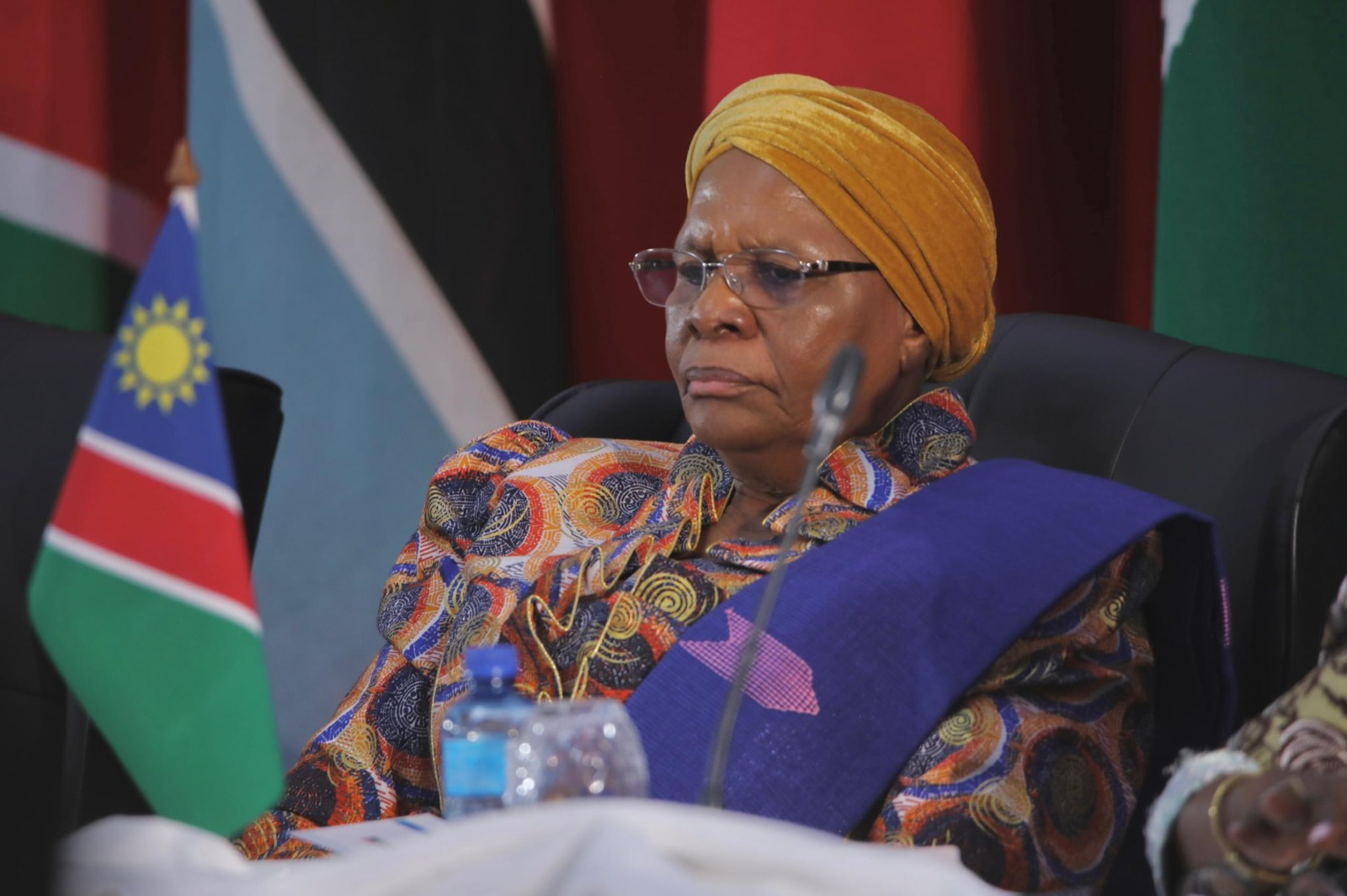Portrait of an elderly woman wearing a yellow head wrap and a colorful patterned dress, seated at a conference with the Namibian flag in the background.