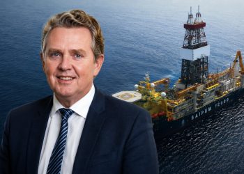 Smiling businessman in a dark suit with a striped tie, set against an offshore oil drilling vessel at sea.