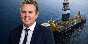 Smiling businessman in a dark suit with a striped tie, set against an offshore oil drilling vessel at sea.