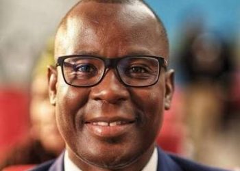 Portrait of a smiling Black man wearing glasses and a navy suit, in a blurred indoor setting.