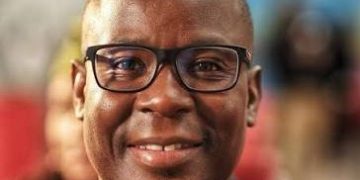 Portrait of a smiling Black man wearing glasses and a navy suit, in a blurred indoor setting.