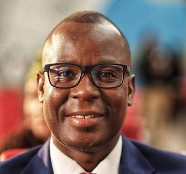 Portrait of a smiling Black man wearing glasses and a navy suit, in a blurred indoor setting.